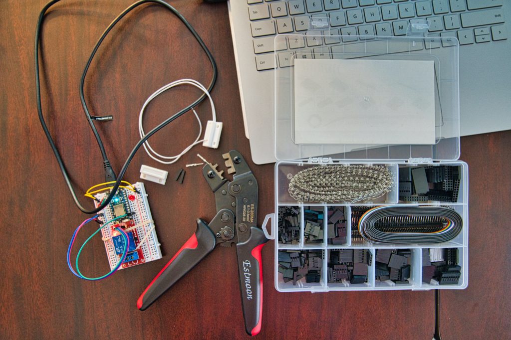 A wire crimper, particle IOT device, magnetic reed switch, laptop, and jumper wires at the start of an internet of things connected project