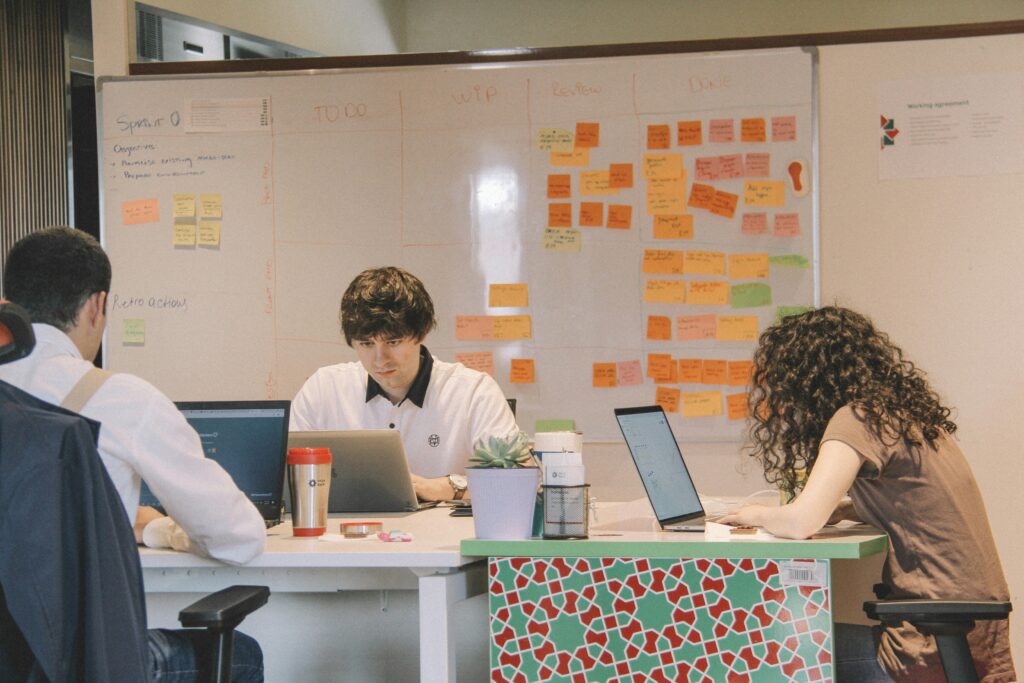 Team members working on laptops in front of a project management board filled with sticky notes, organized by task progress stages.